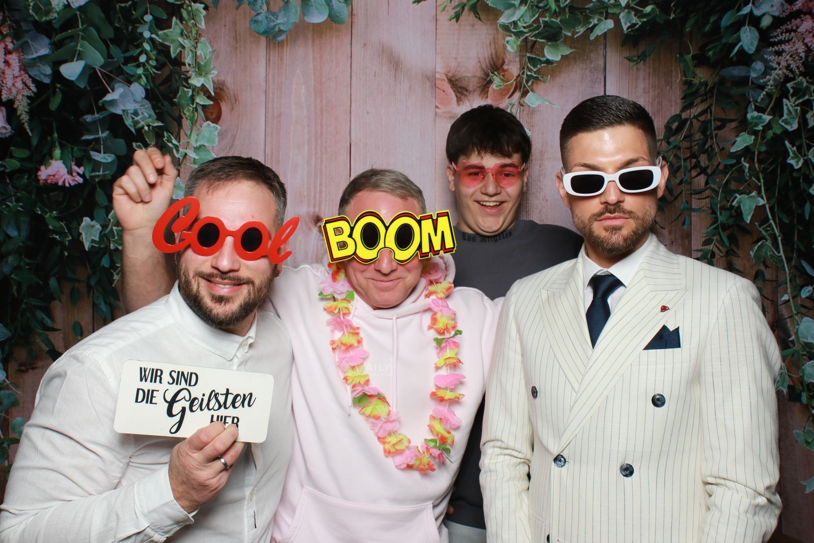 Vier mannen met feestbrillen en bloemenkrans voor een bloemenwand